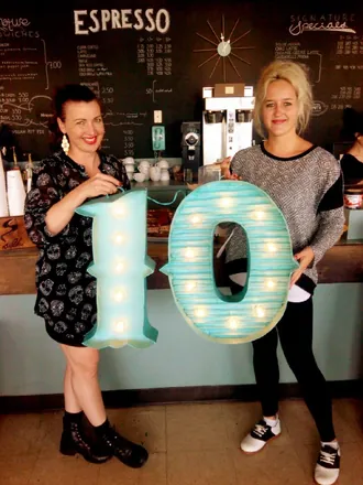 Two women in a coffee shop holding up a giant lit-up number 10