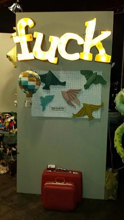 A marquee-light sign spelling a swear word, above a pegboard of paper birds